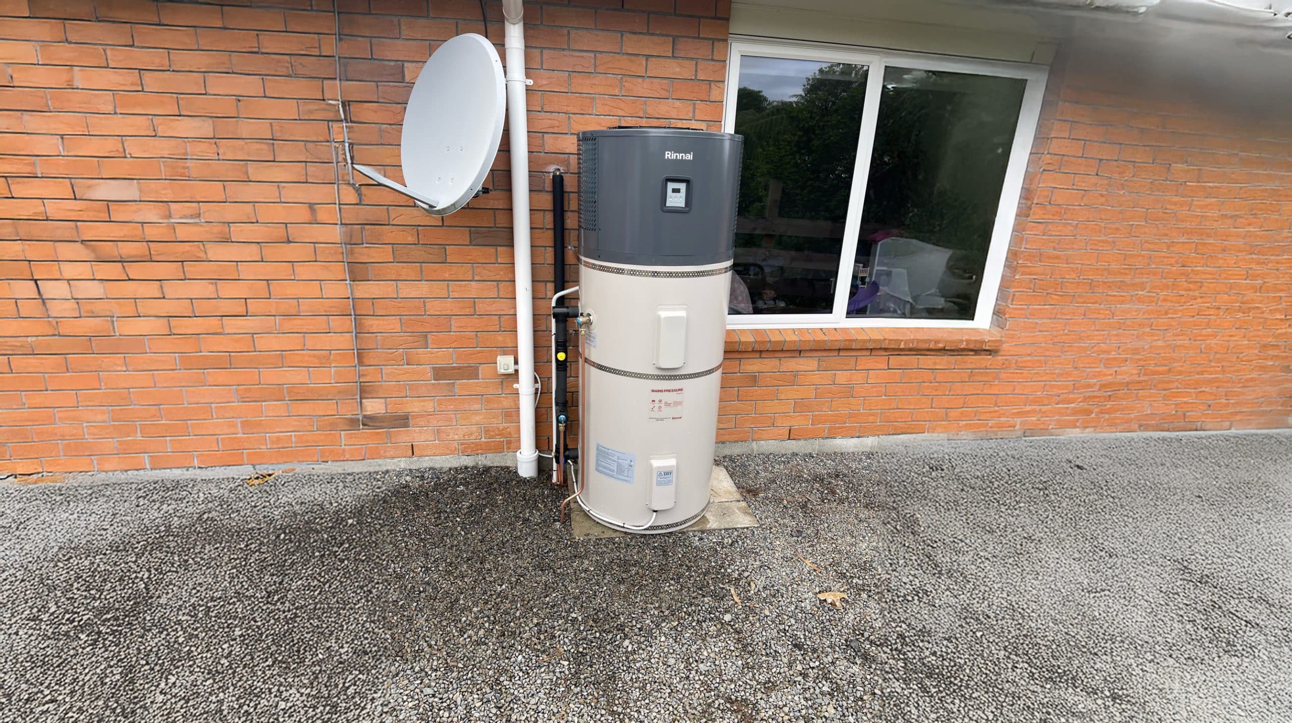 A two-piece Rinnai hot water heat pump system is installed next to a brick building. The unit consists of a tall, cylindrical tank and a smaller, cylindrical component on top. A satellite dish is mounted on the wall above the hot water heat pump.