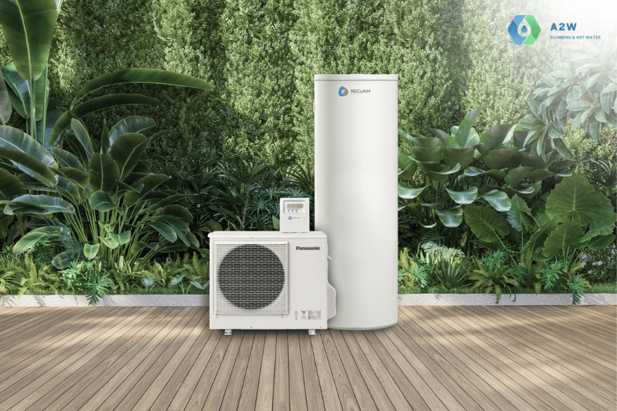 A modern, white Panasonic hot water heat pump system, consisting of an outdoor condenser unit and a tall, cylindrical water storage tank, is shown installed on a wooden deck. The entire system is set against a lush green wall of various plants and foliage. A small logo for A2W is visible in the top right corner.